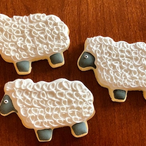May include: Three white sugar cookies decorated to look like sheep. Each cookie has a gray face with a black eye and gray legs. The cookies are on a brown wooden surface.