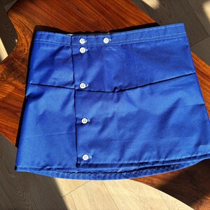 May include: A vibrant blue skirt with white buttons. The skirt is made of a smooth fabric and features a wrap-style design. The buttons are evenly spaced and add a classic touch to the garment. The skirt is displayed on a wooden surface.