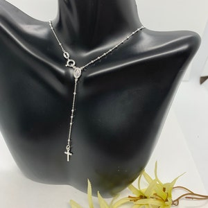 May include: A silver rosary necklace with a small cross charm and a pendant featuring the Virgin Mary. The necklace is displayed on a black mannequin bust.