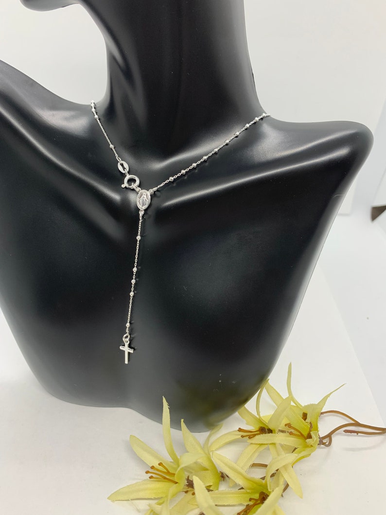 May include: A silver rosary necklace with a small cross charm and a pendant featuring the Virgin Mary. The necklace is displayed on a black mannequin bust.