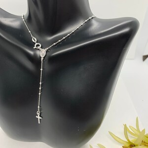 May include: A silver rosary necklace with a small cross and a charm of the Virgin Mary. The necklace is on a black mannequin.
