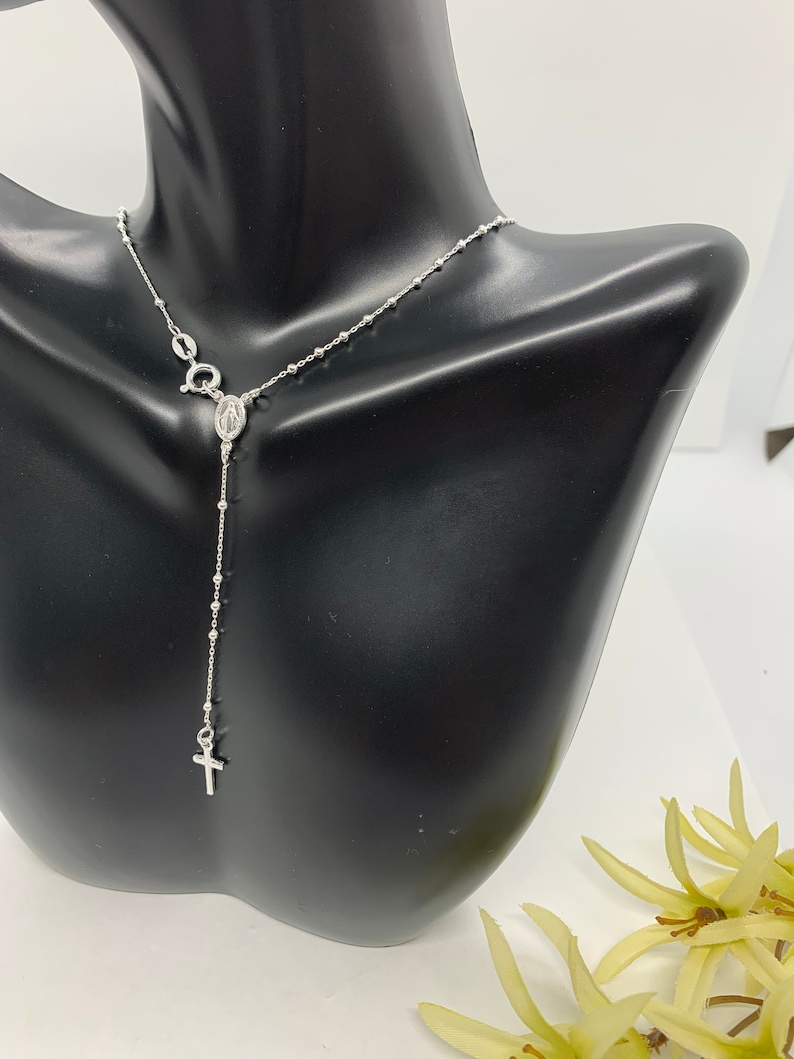 May include: A silver rosary necklace with a small cross and a charm of the Virgin Mary. The necklace is on a black mannequin.