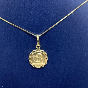 Puede incluir: Un collar de cadena de oro con un colgante que presenta un peque&ntilde;o &aacute;ngel con alas. El colgante es redondo y tiene un dise&ntilde;o detallado.