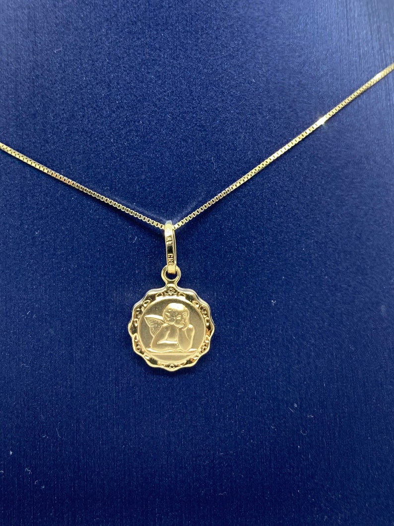 Puede incluir: Un collar de cadena de oro con un colgante que presenta un peque&ntilde;o &aacute;ngel con alas. El colgante es redondo y tiene un dise&ntilde;o detallado.