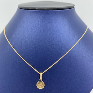 Op de afbeelding: Een gouden ketting met een klein rond hanger met een engelontwerp.