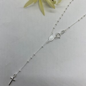 May include: A silver chain necklace with a small cross pendant and a silver oval charm. The chain is made of small beads.