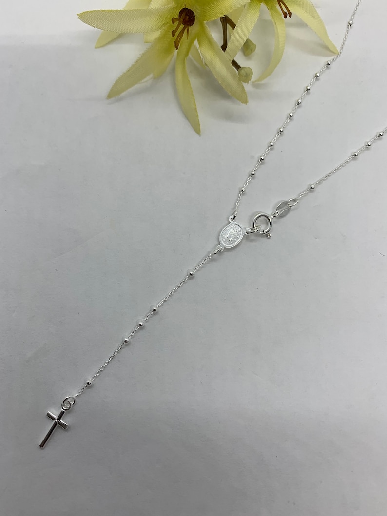 May include: A silver chain necklace with a small cross pendant and a silver oval charm. The chain is made of small beads.