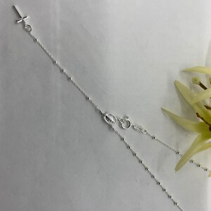 May include: A silver chain necklace with a small cross pendant and a charm of the Virgin Mary. The chain is made of small, round beads.
