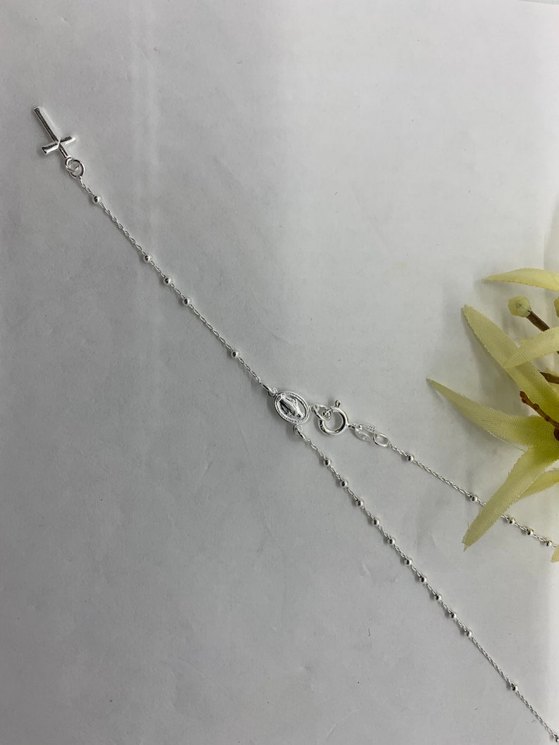 May include: A silver chain necklace with a small cross pendant and a charm of the Virgin Mary. The chain is made of small, round beads.