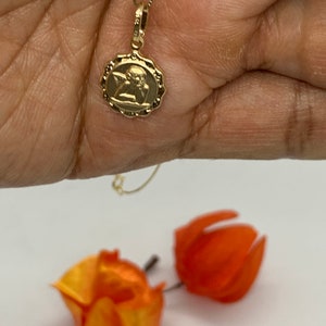 Puede incluir: Un collar de cadena de oro con un peque&ntilde;o colgante redondo que presenta una imagen detallada de un &aacute;ngel. El colgante tiene un borde festoneado.