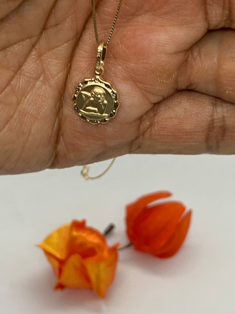 Puede incluir: Un collar de cadena de oro con un peque&ntilde;o colgante redondo que presenta una imagen detallada de un &aacute;ngel. El colgante tiene un borde festoneado.