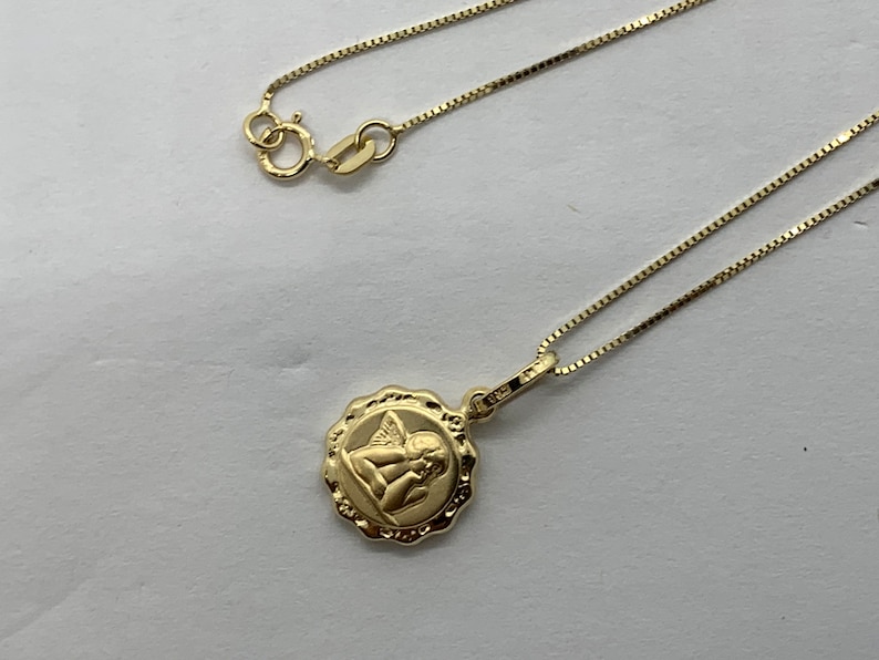 Puede incluir: Un collar de cadena de oro con un colgante redondo que presenta una imagen detallada de un &aacute;ngel con alas.