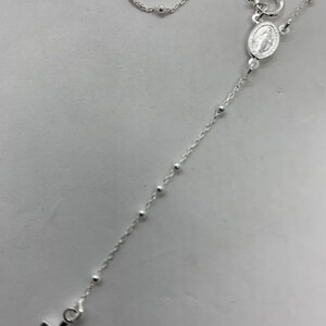 May include: A silver chain necklace with a small cross pendant and a silver charm with a religious image. The chain is made of small, round beads.