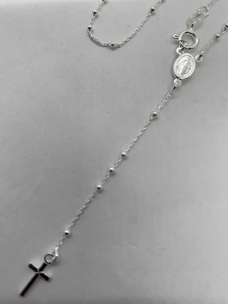 May include: A silver chain necklace with a small cross pendant and a silver charm with a religious image. The chain is made of small, round beads.