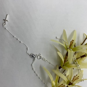 May include: A silver rosary necklace with a small cross charm and a silver medal charm. The necklace is on a white background with a yellow flower.