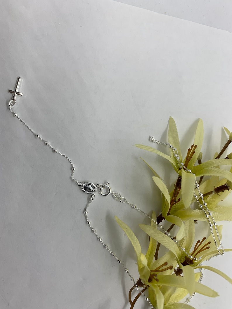 May include: A silver rosary necklace with a small cross charm and a silver medal charm. The necklace is on a white background with a yellow flower.
