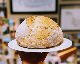 May include: A freshly baked loaf of sourdough bread on a white plate. The bread has a golden brown crust and a soft, fluffy interior.