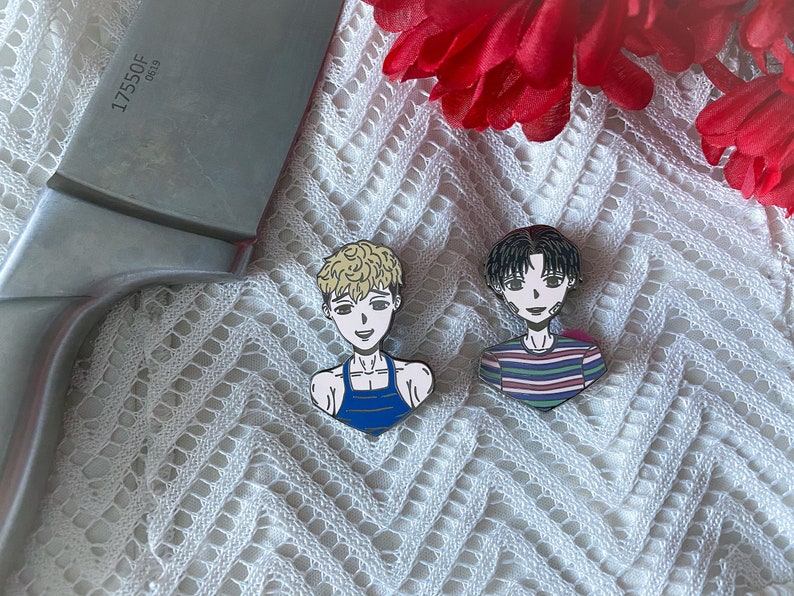 Puede incluir: Dos pines de esmalte con personajes de anime. El pin de la izquierda presenta un personaje rubio con un tanque azul. El pin de la derecha presenta un personaje de cabello negro con una camisa a rayas.