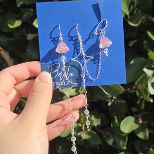 May include: Three pairs of earrings on a blue card. The earrings feature pink flower-shaped pendants, clear beads, and silver chains. One pair has a star design. The card has the text "Bella Jewelry".