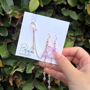 May include: A pair of gold-colored earrings with dangling clear beads and light purple flower-shaped accents. The earrings are connected by a delicate chain to a gold-colored ear cuff. The earrings are displayed on a white card with the "Belafas Jewelry" logo.