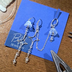 May include: A pair of silver earrings with a star design and dangling clear beads. Each earring features a light blue flower-shaped charm. The earrings are connected by a delicate chain. A blue card with the text "Bella Ray Jewelry" is in the background.