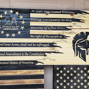 Op de afbeelding: Een houten bord met een zwart-wit Amerikaans vlagontwerp. Het bord bevat de tekst "2A Protect the 2nd Amendment" en de tekst van het Tweede Amendement op de Grondwet van de Verenigde Staten. Het bord bevat ook een Spartaanse helm met de Griekse woorden "ΜΟΛΩΝ ΛΑΒΕ" (Molon Labe) wat "Kom en haal ze" betekent.