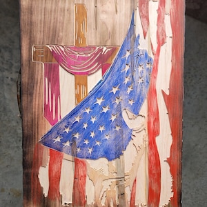 May include: A wooden sign with a distressed finish featuring a red, white, and blue American flag draped over a wooden cross. The flag is partially covering the cross, and the cross is decorated with a purple and pink fabric drape. The sign is a patriotic and religious themed piece of art.