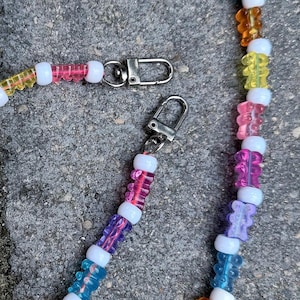 May include: A beaded lanyard featuring translucent, multi-colored gummy bear-shaped charms alternating with opaque white spherical beads. The accessory includes two silver-toned metal lobster claw clasps attached to the ends of the beaded strand.