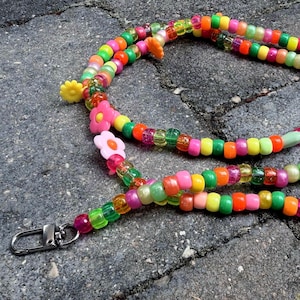 May include: A lanyard made of multi-colored plastic beads in shades of pink, orange, yellow, and green. The strand features several flower-shaped beads and is finished with two silver-toned metal lobster claw clasps at each end of the cord.