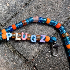 May include: A beaded lanyard featuring translucent blue, glittery orange, and clear beads strung on a blue cord. The word PLUGZ is spelled out with individual letter beads in light blue, white, pink, and purple. A silver metal lobster clasp is attached.