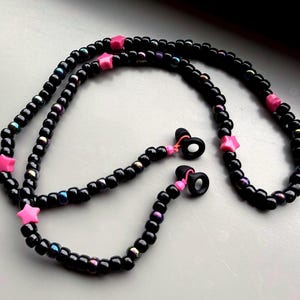 May include: Black beaded eyeglass chain with pink star accents. The chain features small, iridescent beads and pink star-shaped beads. Includes black earbud holders with pink accents.