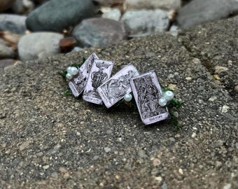 May include: A collection of tarot cards with black and white illustrations and text, including "ACE OF CUPS" and "THE POPE". The cards are arranged on a bed of greenery and pearls, set against a textured stone background.