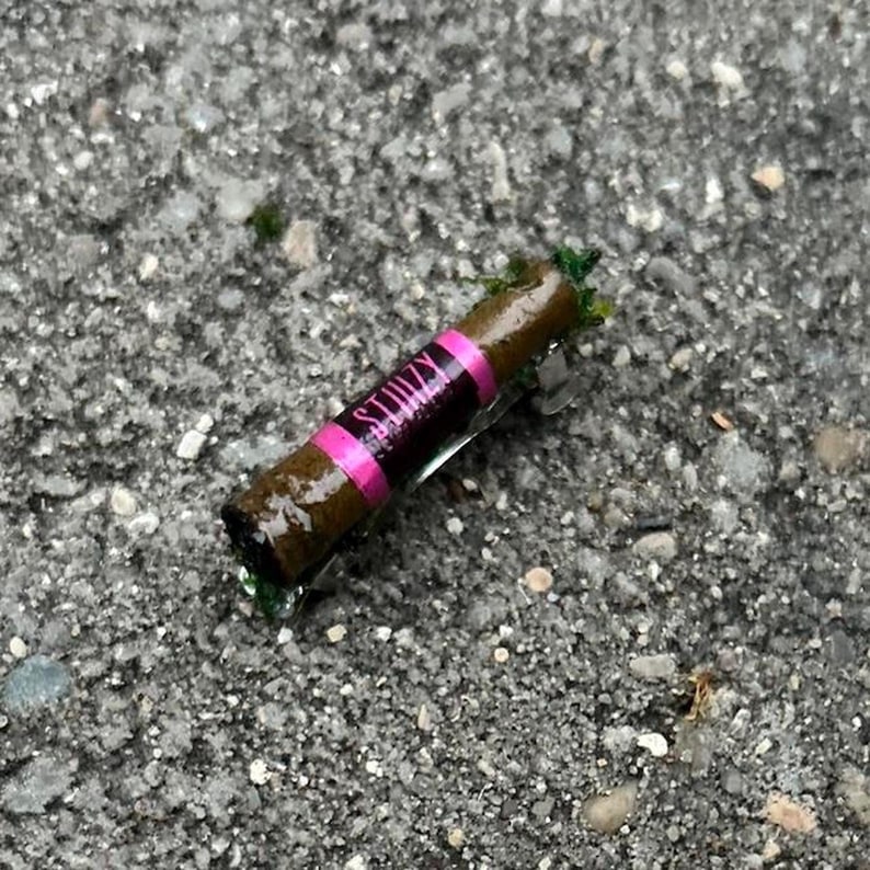 Puede incluir: Un producto enrollado marr&oacute;n y verde con una etiqueta rosa y negra que dice "STIIIZY". El producto est&aacute; sobre una superficie gris y texturizada.