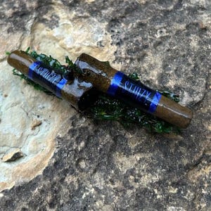 May include: Two brown wooden objects with blue bands and the word "STIIIZY" printed on them. The objects are resting on a bed of green moss, set against a textured stone background.