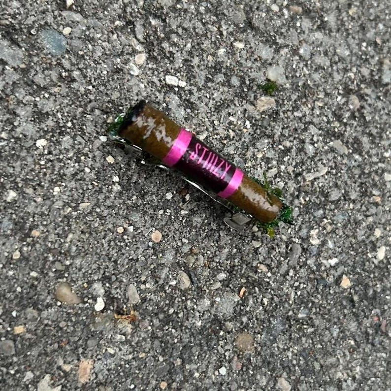 Puede incluir: Un producto enrollado marr&oacute;n con una banda rosa que dice "STIIIZY" en letras blancas. El producto descansa sobre una superficie gris texturizada. El extremo del producto es negro.