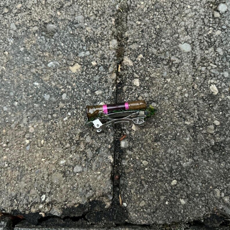 Puede incluir: Un peque&ntilde;o tap&oacute;n de botella marr&oacute;n con bandas rosas y un abridor de botellas plateado. El tap&oacute;n descansa sobre una superficie gris texturizada.