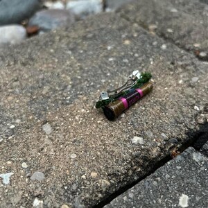 Puede incluir: Una peque&ntilde;a pinza para el cabello con un dise&ntilde;o cil&iacute;ndrico marr&oacute;n y rosa, adornada con una franja rosa y la palabra "CTUZY". Cuenta con un cierre de metal plateado y est&aacute; decorada con elementos verdes.
