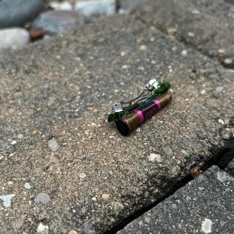 Puede incluir: Una peque&ntilde;a pinza para el cabello con un dise&ntilde;o cil&iacute;ndrico marr&oacute;n y rosa, adornada con una franja rosa y la palabra "CTUZY". Cuenta con un cierre de metal plateado y est&aacute; decorada con elementos verdes.