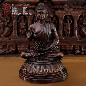 May include: A bronze Buddha statue in a seated meditation pose with one hand raised in the Abhaya mudra gesture. The statue is 22.5 inches tall and weighs 15.960 pounds. The statue is sitting on a round base with intricate carvings.