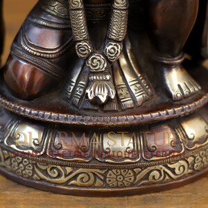 May include: A bronze statue of a deity with intricate details, including a beaded necklace and a patterned skirt. The statue is sitting on a round base with a silver-colored band and ornate carvings.