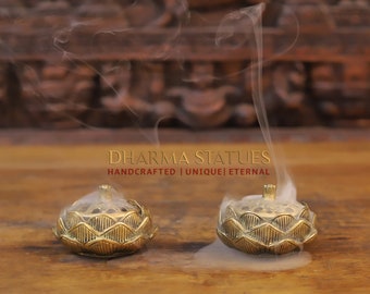 May include: Two gold lotus incense burners with smoke rising from them. The burners are on a wooden surface. The text "DHARMA STATUES HANDCRAFTED | UNIQUE | ETERNAL" is visible in the image.