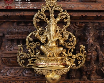 Könnte beinhalten: Eine goldfarbene Wanddekoration aus Messing mit einer detaillierten Darstellung der hinduistischen Gottheit Ganesha. Das komplizierte Design umfasst verzierte Schnörkel und eine kleine Öllampe. Die Wanddekoration ist 35,56 cm hoch, 27,94 cm lang und 19,05 cm breit. Sie wiegt 3,760 Kilogramm.