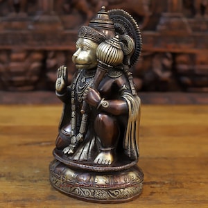 May include: A brass statue of the Hindu deity Hanuman, depicted in a seated pose with his right hand raised in a gesture of blessing. The statue is finished in a combination of gold and copper tones, with intricate details throughout. The statue is sitting on a circular base with a decorative design.