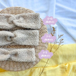 Op de afbeelding: Drie beige, donzige haarbanden worden gepresenteerd op een gevlochten mand. De haarbanden zijn gestapeld, met wolk-vormige labels die leeftijdsgroepen aangeven: 1-2, 3-5 en 6-12 jaar. De achtergrond omvat witte en gele stof.