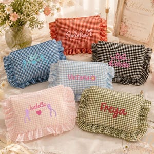 May include: Six rectangular zippered pouches with ruffled edges, each featuring a unique gingham pattern in red, blue, black, light blue, pink, or green. Each pouch is embroidered with a name and decorative icons like bows, hearts, or flowers.