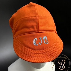 Pode incluir: Um boné laranja brilhante com uma coroa estruturada e uma aba curva com costuras decorativas. Na parte frontal da aba, as letras CJQ estão bordadas numa fonte gótica azul-clara, criando um contraste nítido com o tecido laranja.
