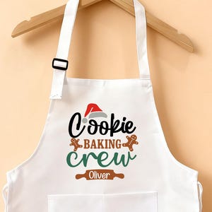 May include: White apron with the text "Cookie Baking Crew" in black, green, and brown. The apron has a pocket and adjustable neck strap. The apron is hanging on a wooden hanger.