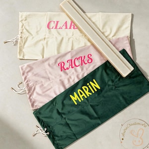 May include: Three rectangular fabric storage bags with drawstring closures, featuring the text CLAR, RACKS, and MARIN in pink, hot pink, and yellow. A long, narrow, light-coloured wooden bar rests diagonally across the top of the bags.