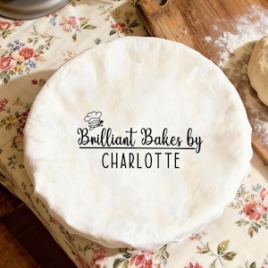 Op de afbeelding: Een witte deegafdekking met de tekst "Brilliant Bakes by CHARLOTTE" in zwart schrift. Een deegbal en een houten snijplank bestrooid met bloem zijn ook zichtbaar. De scène speelt zich af op een tafelkleed met bloemenpatroon.