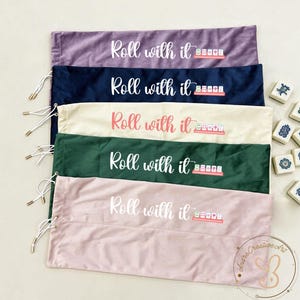 May include: Five rectangular fabric drawstring bags in purple, navy blue, cream, dark green, and light pink. Each bag features the white or pink text Roll with it and a graphic of five mahjong tiles on a rack. Small mahjong tiles are nearby.